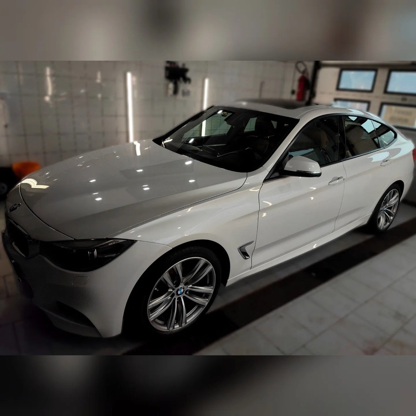 Białe BMW serii 3 Gran Turismo po umyciu, widoczne odbicia światła na lakierze, w tle ściana myjni z oświetleniem i sprzętem.