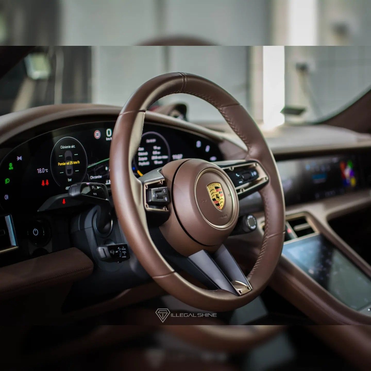 Wnętrze samochodu Porsche Taycan z brązową skórzaną kierownicą, widoczne cyfrowe zegary i centralny ekran dotykowy, logo firmy na kierownicy, dolny napis 'ILLEGAL SHINE'.