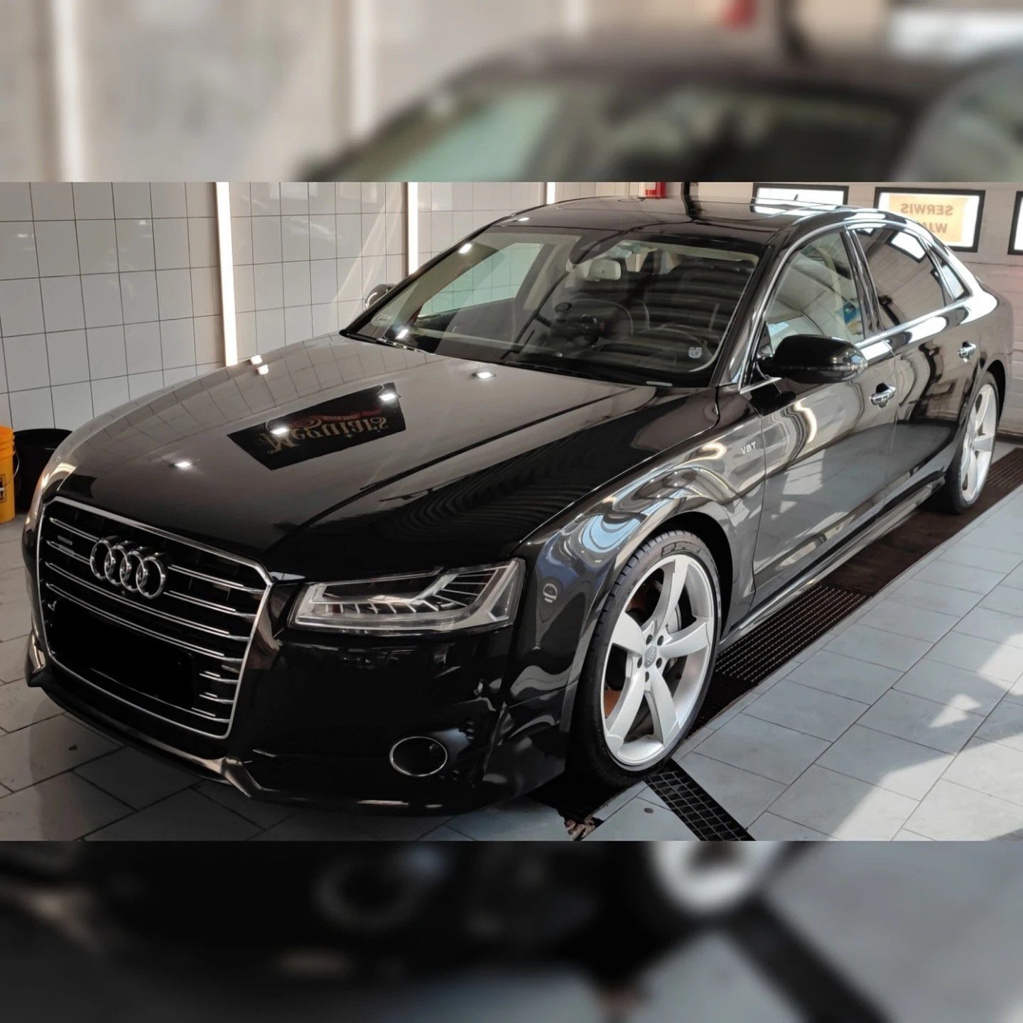 Błyszczący, czarny samochód Audi A8 po umyciu, zaparkowany w jasnym pomieszczeniu z białymi kafelkami i kratką odpływową w podłodze.