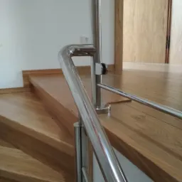 Polerowana, metalowa poręcz zamontowana na drewnianych schodach wewnątrz domu, widok z dołu, drzwi z jasnego drewna w tle.