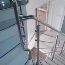 Polerowana, metalowa balustrada schodowa z poziomymi prętami, zamontowana na schodach z szarymi paskami antypoślizgowymi, widok z góry w dół, w tle widoczne drzwi.