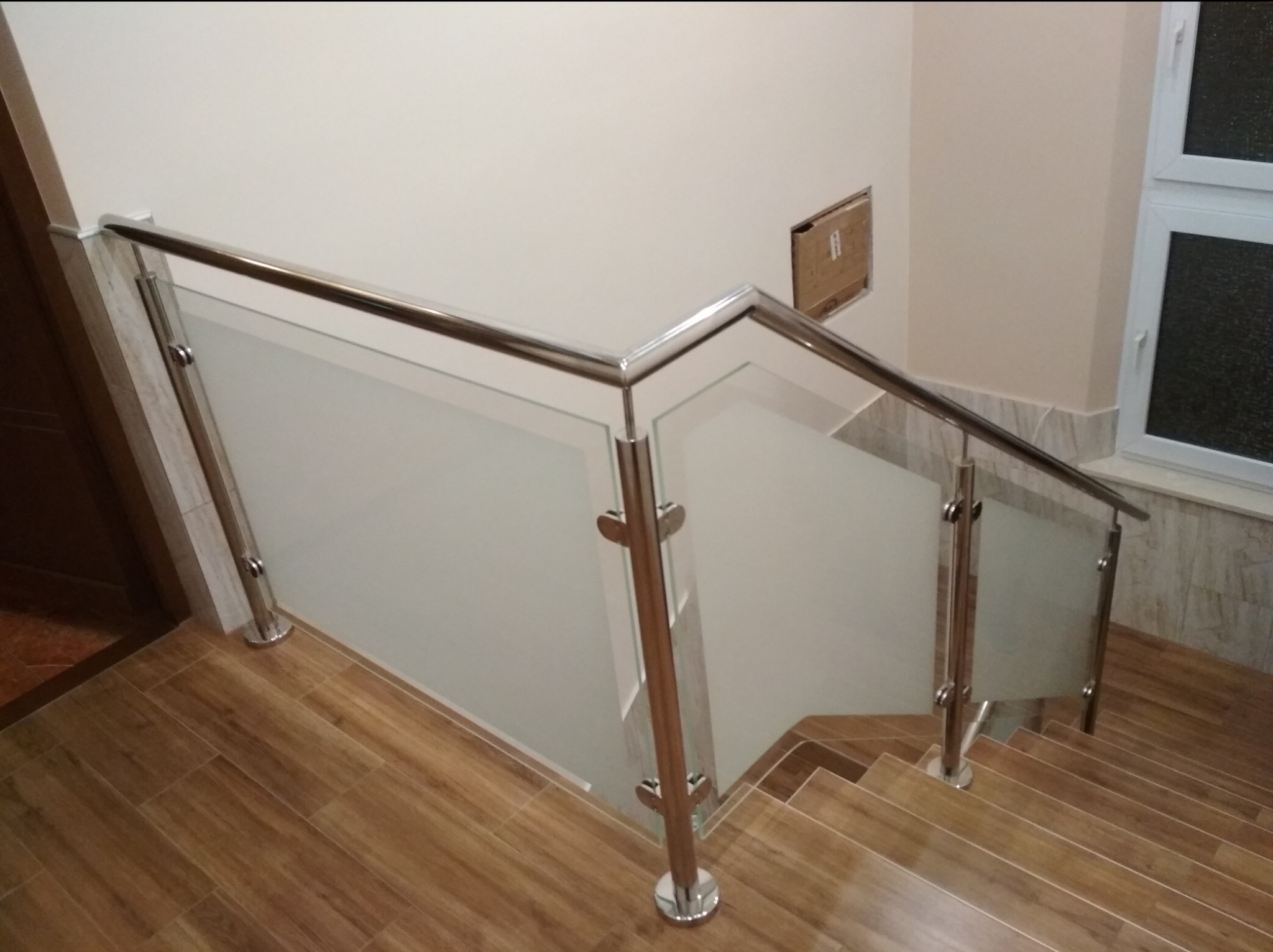 Szklana balustrada z elementami ze stali nierdzewnej zamontowana na schodach wyłożonych płytkami imitującymi drewno. Widoczny fragment okna po prawej stronie.