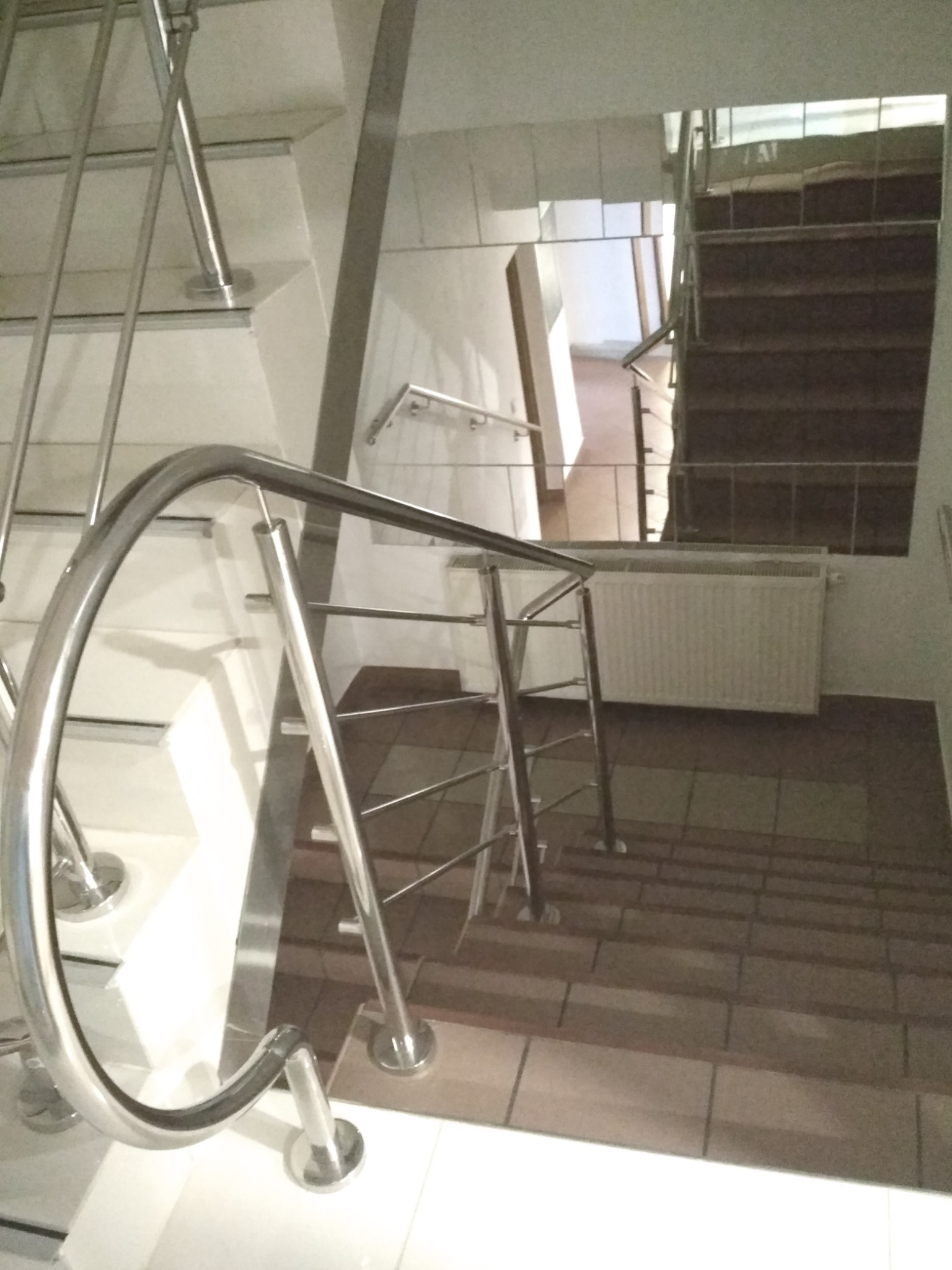 Klatka schodowa z metalową poręczą i schodami wyłożonymi brązowymi płytkami, widok z góry na dół.