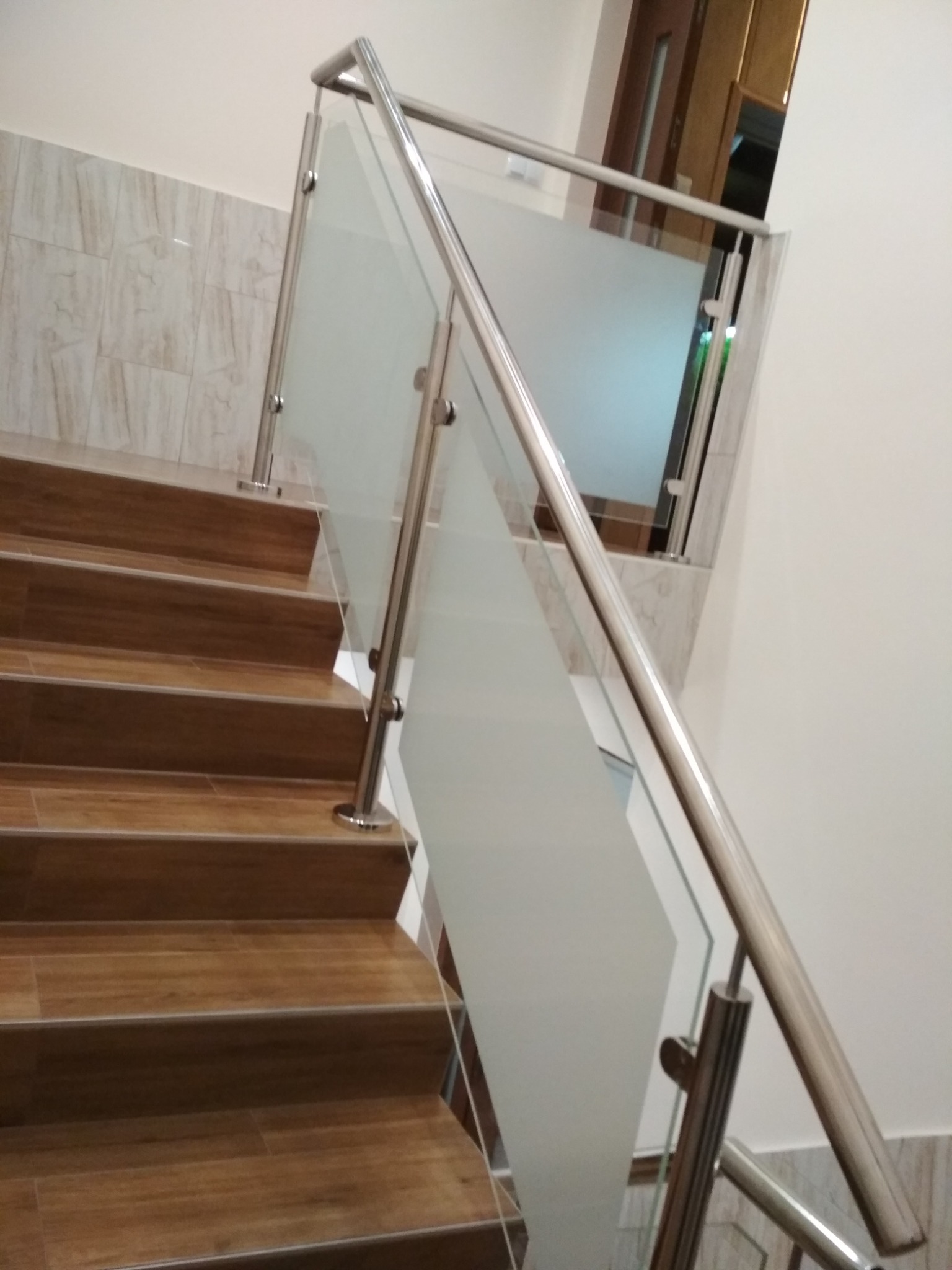 Schody z okładziną drewnopodobną, zabezpieczone szklaną balustradą z nierdzewnymi poręczami, widoczne z dołu pod kątem.