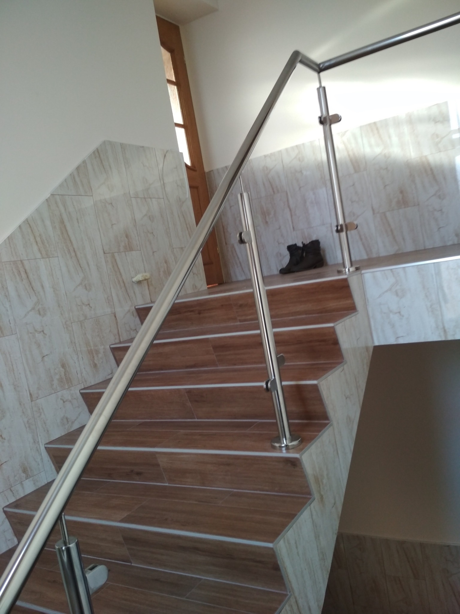 Wnętrze domu z drewnopodobnymi schodami i błyszczącą, metalową balustradą; na górnym biegu schodów para ciemnych butów.