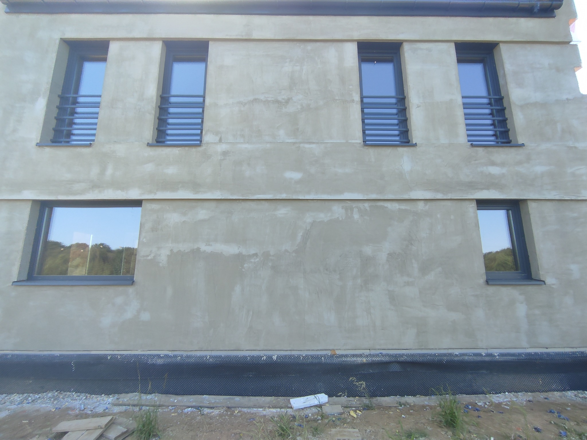 Elewacja budynku w trakcie wykańczania z zamontowanymi oknami i metalowymi balustradami okiennymi, widoczna warstwa hydroizolacji przy gruncie.