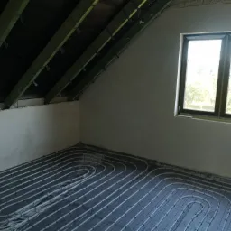 Poddasze z oknem, na podłodze rozłożone rury ogrzewania podłogowego na czarnej macie izolacyjnej, ściany przygotowane do dalszych prac wykończeniowych.