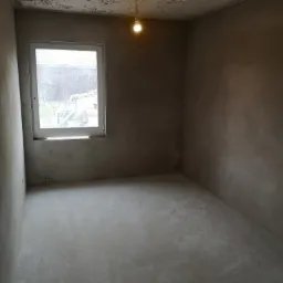 Surowe wnętrze pokoju po tynkowaniu maszynowym, widoczne świeże tynki na ścianach i suficie z miejscowymi ubytkami, plastikowe okno z widokiem na zewnątrz, pojedyncza żarówka oświetla pomieszczenie.