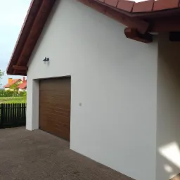 Świeżo otynkowana, biała elewacja garażu z brązową bramą segmentową i czerwoną dachówką, widok z zewnątrz, kostka brukowa na podjeździe.