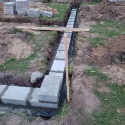 Wykopany fundament z ułożonymi bloczkami betonowymi na zaprawie, widoczne sznurki wyznaczające linię, drewniane stemple i przygotowane bloczki w tle.