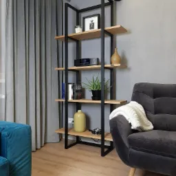Regał z drewnianymi półkami i czarną metalową konstrukcją, ustawiony w salonie z szarą ścianą, obok szarego fotela z narzutą i fragmentu turkusowej sofy.