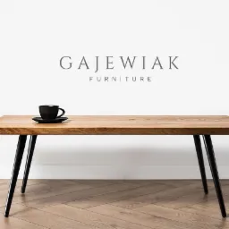 Stylowy stolik kawowy z blatem z jasnego drewna i czarnymi, stożkowymi nogami, na blacie czarna filiżanka z kawą, na tle białej ściany i podłogi ułożonej w jodełkę, z widocznym logo producenta mebli.