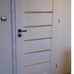 Białe drzwi wewnętrzne z poziomymi przeszkleniami, czarna klamka, widoczne zawiasy, zamontowane w jasnym pomieszczeniu z panelami podłogowymi.