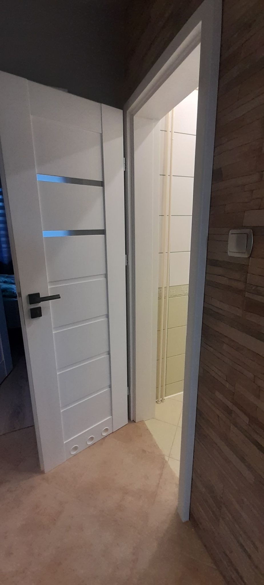 Otwarta, biała, nowoczesna konstrukcja z poziomymi przeszkleniami i czarną klamką, widok na łazienkę z jasnymi płytkami i fragment ściany z okładziną imitującą drewno.