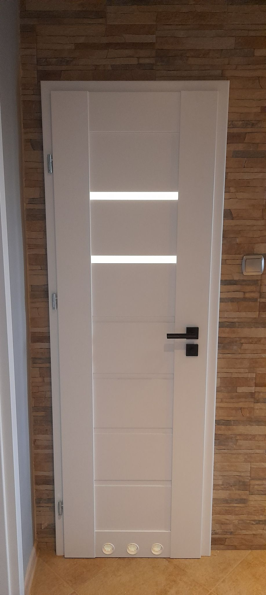 Białe drzwi wewnętrzne z trzema poziomymi przeszkleniami, czarną klamką i oświetleniem podłogowym, zamontowane w ścianie z dekoracyjnej cegły.