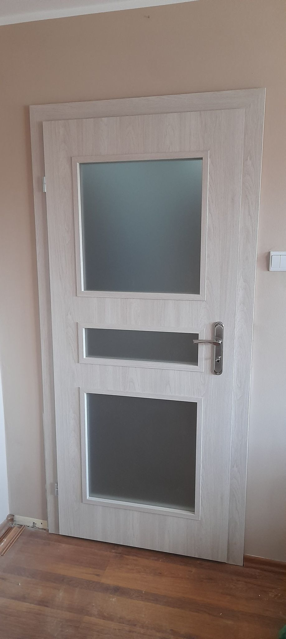 Jasne, drewnopodobne drzwi wewnętrzne z trzema przeszkleniami zamontowane w mieszkaniu w Pabianicach, widoczne niedokończone wykończenie przy podłodze.