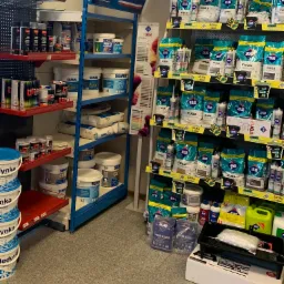 Półki sklepowe wypełnione materiałami budowlanymi, w tym worki z fugą Atlas, wiadra z farbami Semim i pojemniki z innymi produktami do wykończenia wnętrz, z widocznymi logotypami marek.