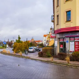 Budynek z szyldem 'Nieruchomości' i plakatami informującymi o kredytach bankowych, widok z mokrej ulicy w pochmurny dzień.