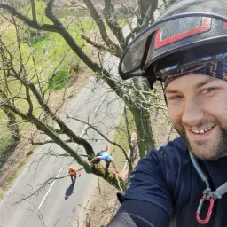 Selfie uśmiechniętego mężczyzny w ochronnym kasku na tle drzewa, z którego inni pracownicy usuwają gałęzie nad jezdnią.