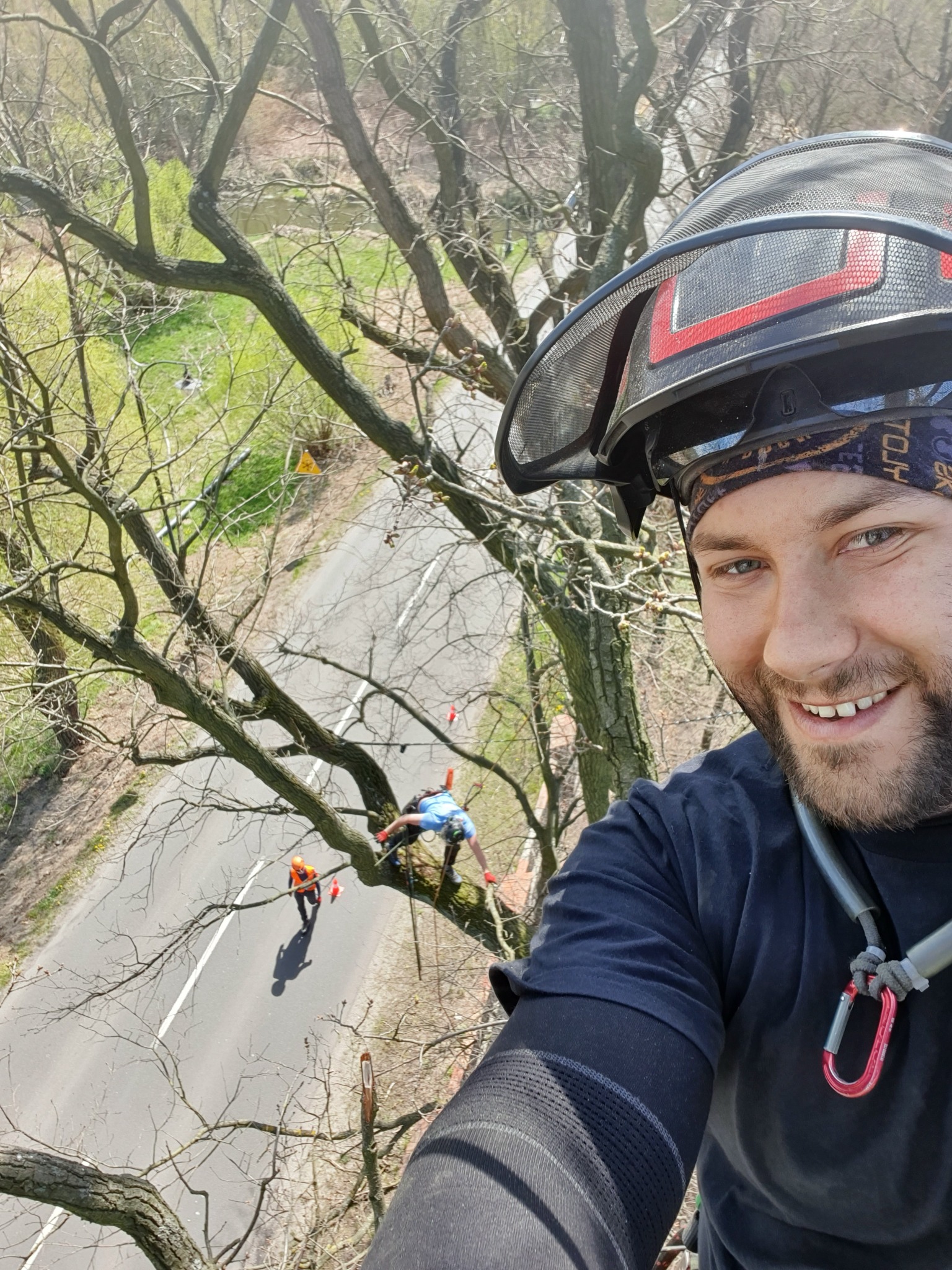 Selfie uśmiechniętego mężczyzny w ochronnym kasku na tle drzewa, z którego inni pracownicy usuwają gałęzie nad jezdnią.