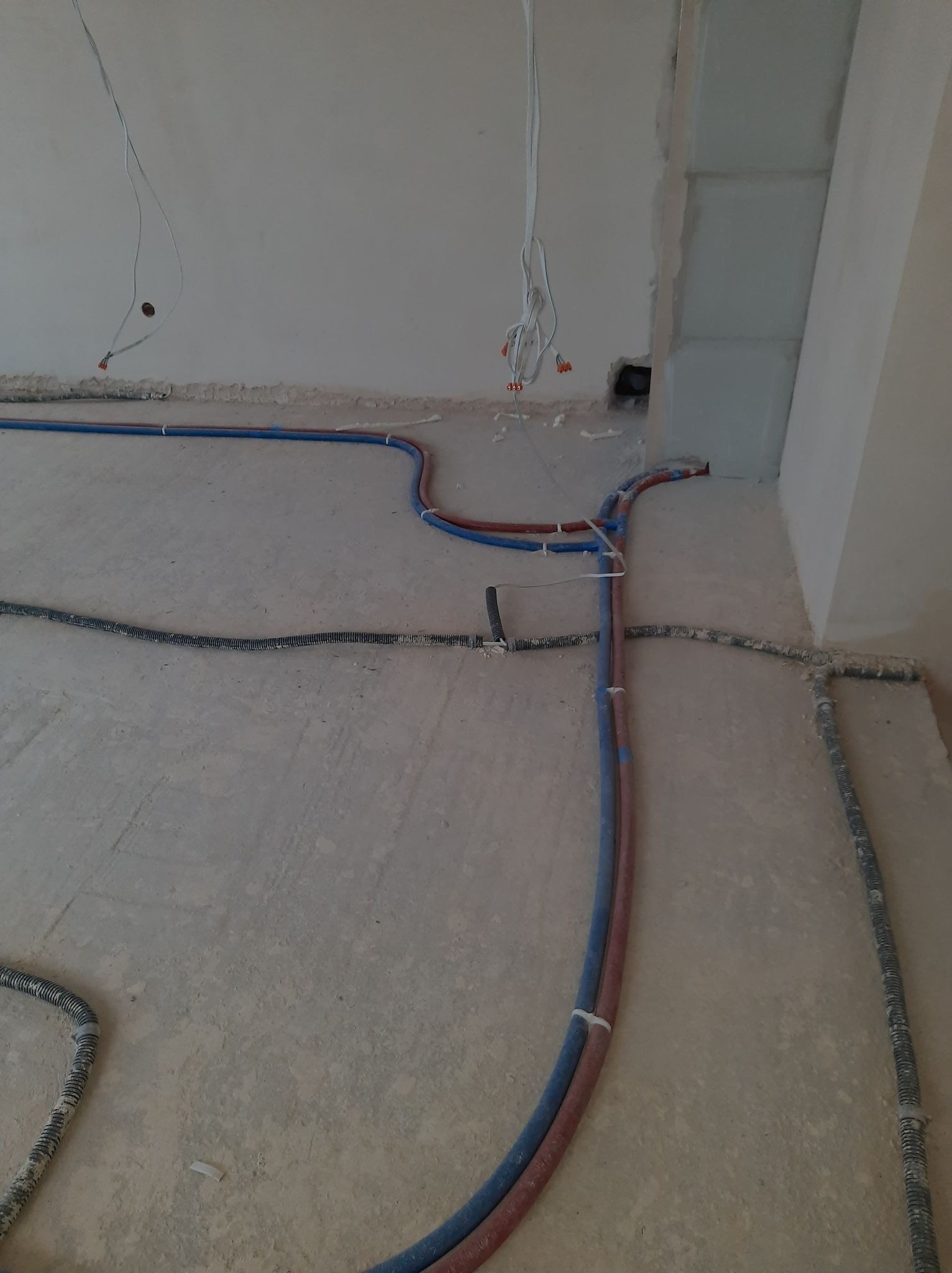 Podłogowe prowadzenie instalacji: rury w kolorach niebieskim i czerwonym, zabezpieczone białymi opaskami, oraz peszel na surowym, betonowym podłożu w trakcie prac wykończeniowych.