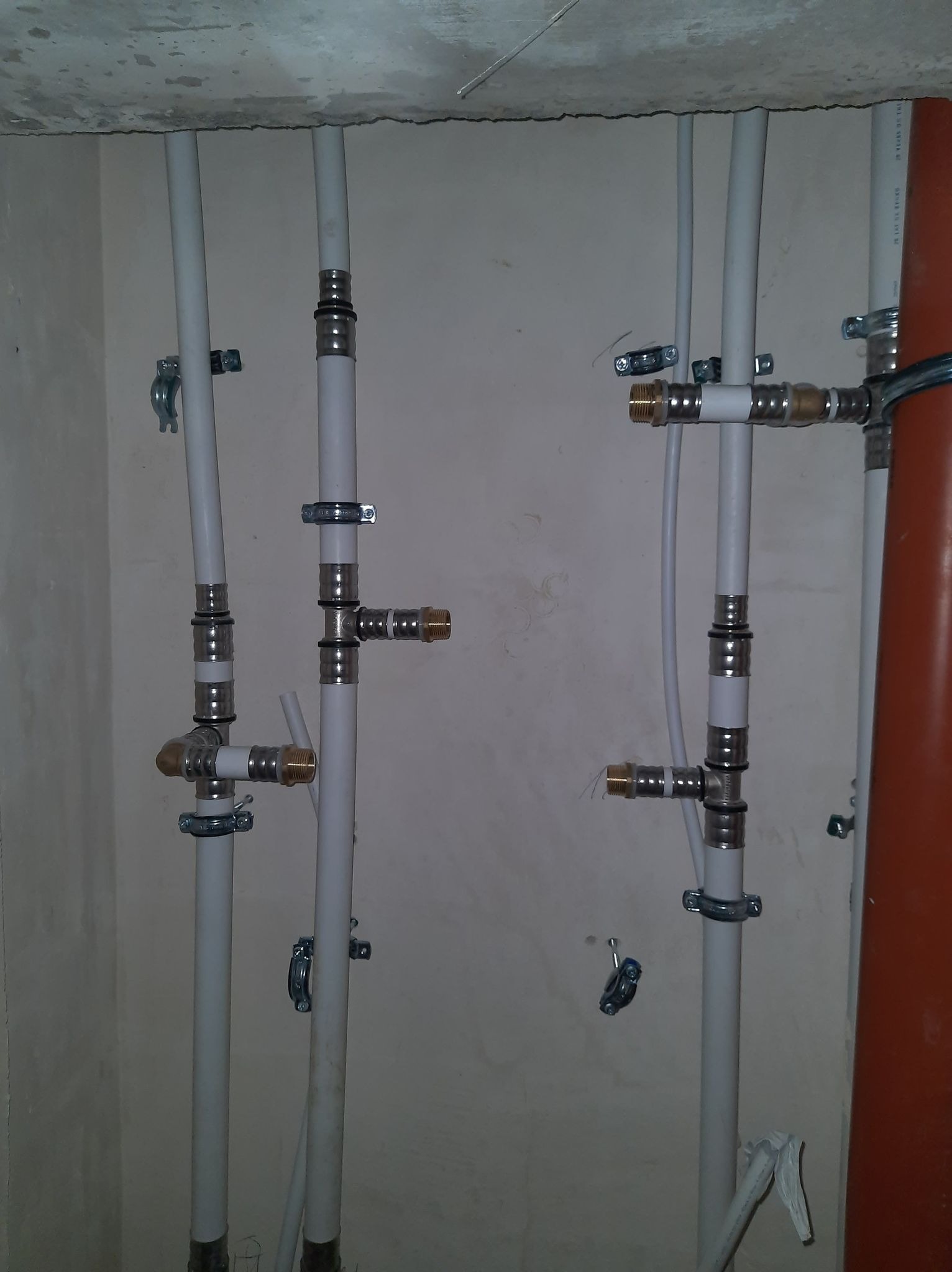Białe rury instalacyjne z metalowymi złączkami i uchwytami mocującymi do ściany, widoczne połączenia typu T i kolanka, surowy stan wykończenia ścian i sufitu.