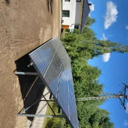 Panel słoneczny ustawiony na metalowej konstrukcji na tle domu, drzew i błękitnego nieba z chmurami, widoczne słupy elektryczne