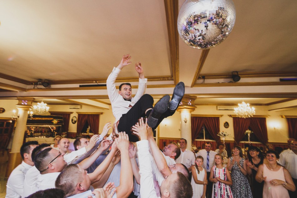 Uśmiechnięty mężczyzna w białej koszuli i czarnych spodniach, podrzucany do góry przez grupę osób na sali balowej z lustrzaną kulą i żyrandolami.