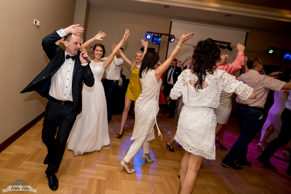 Parkiet weselny w trakcie zabawy: wodzirej z mikrofonem w smokingu, panna młoda w białej sukni i goście weselni tańczą w rytm muzyki, oświetlenie sceniczne w tle.