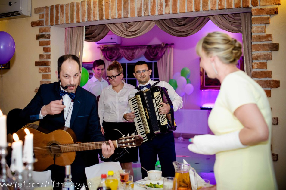 Dwóch muzyków w eleganckich strojach grających na gitarze i akordeonie podczas imprezy, w tle goście i dekoracje balonowe.
