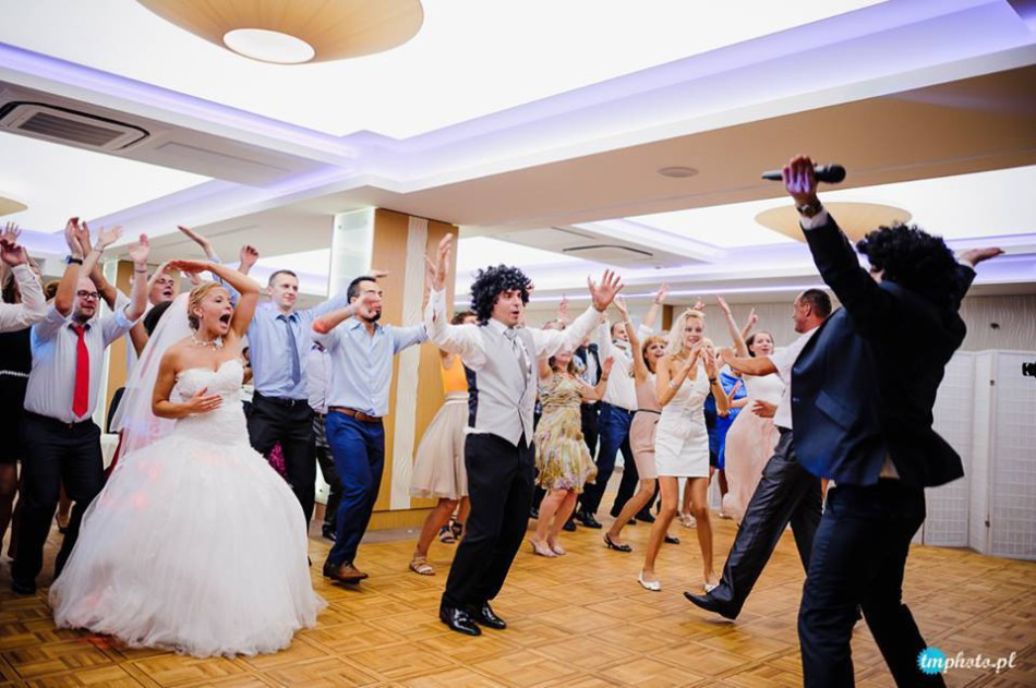 Wnętrze sali balowej podczas wesela: panna młoda w białej sukni, goście i wodzirej w afro peruce tańczą na parkiecie, unosząc ręce w górę.