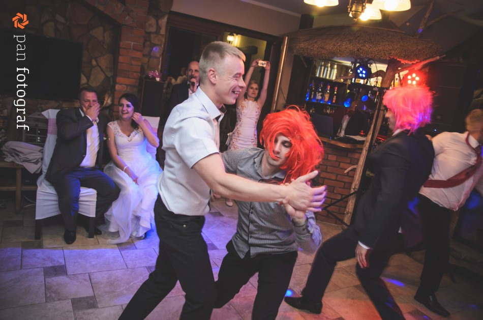 Goście weselni w trakcie zabawy tanecznej, mężczyźni w czerwonych perukach, para młoda obserwująca z boku, bar z oświetleniem w tle.