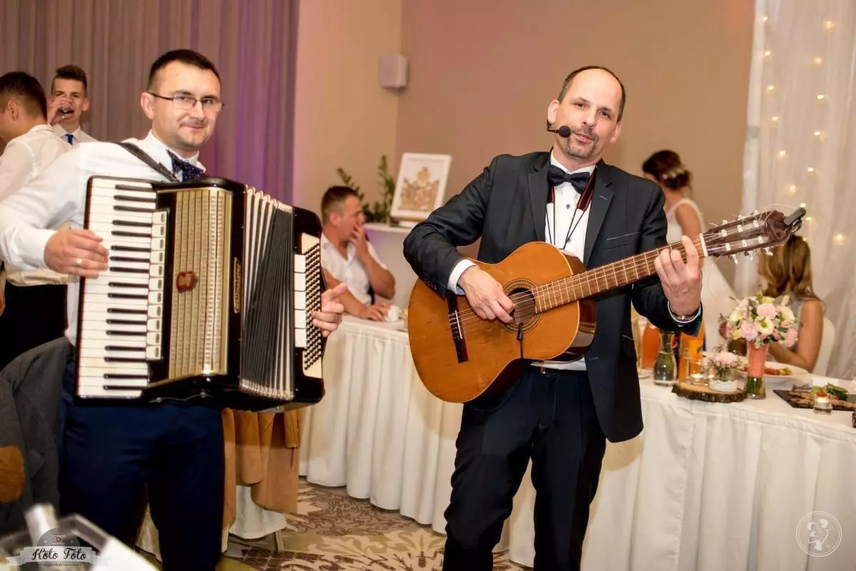 Muzycy w garniturach: akordeonista z muszką i gitarzysta z mikrofonem nagłownym występują na tle sali bankietowej z dekoracjami ślubnymi.