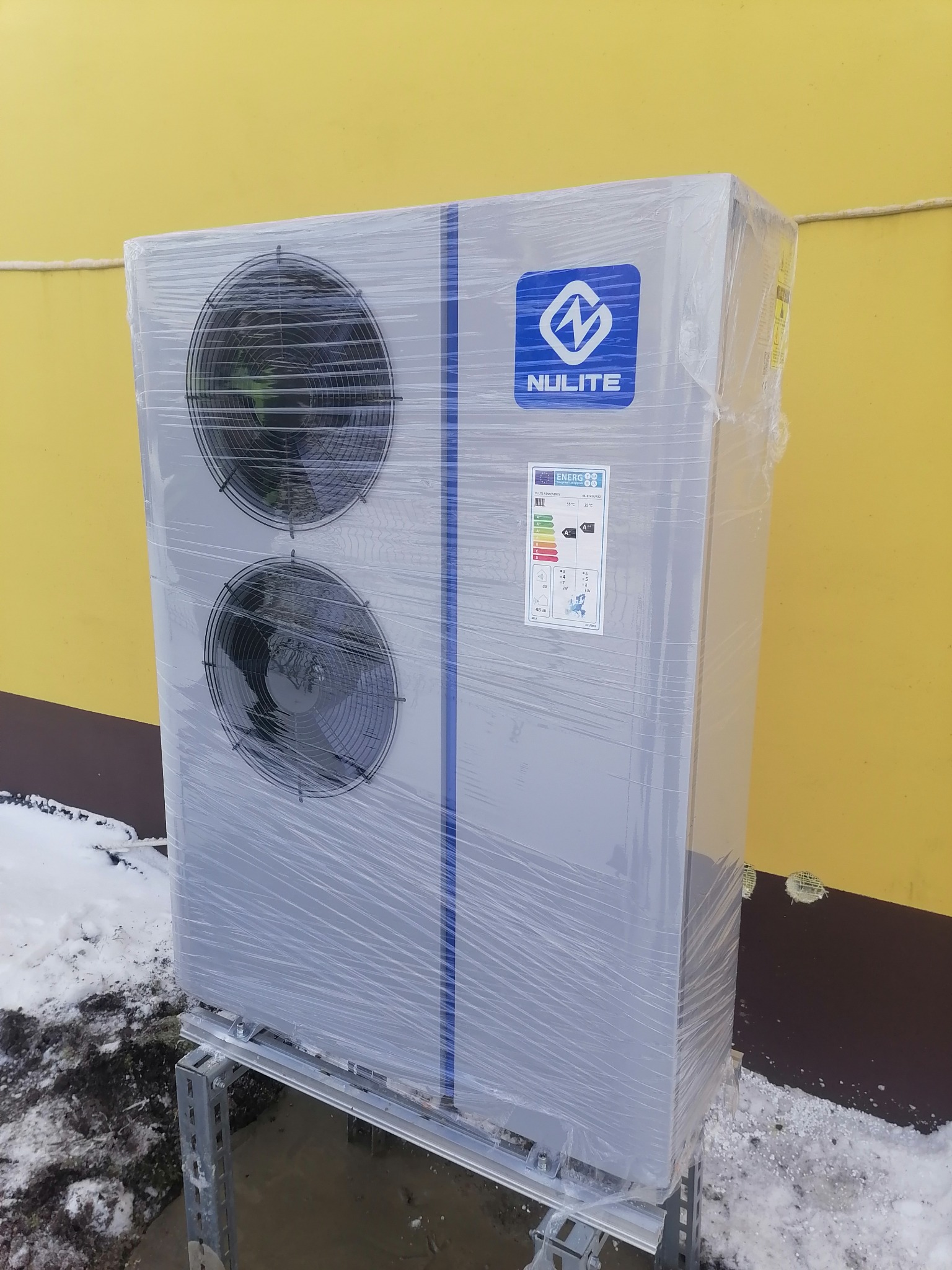 Nowa, owinięta folią pompa ciepła marki Nulite, ustawiona na metalowej konstrukcji, z widocznym fragmentem ośnieżonego terenu i żółtej ściany budynku w tle.