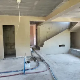 Wnętrze w trakcie prac wykończeniowych: surowy betonowy sufit, ściany pokryte świeżym tynkiem o delikatnym odcieniu beżu, widoczne przewody instalacyjne na podłodze, otwór drzwiowy bez ościeżnicy...