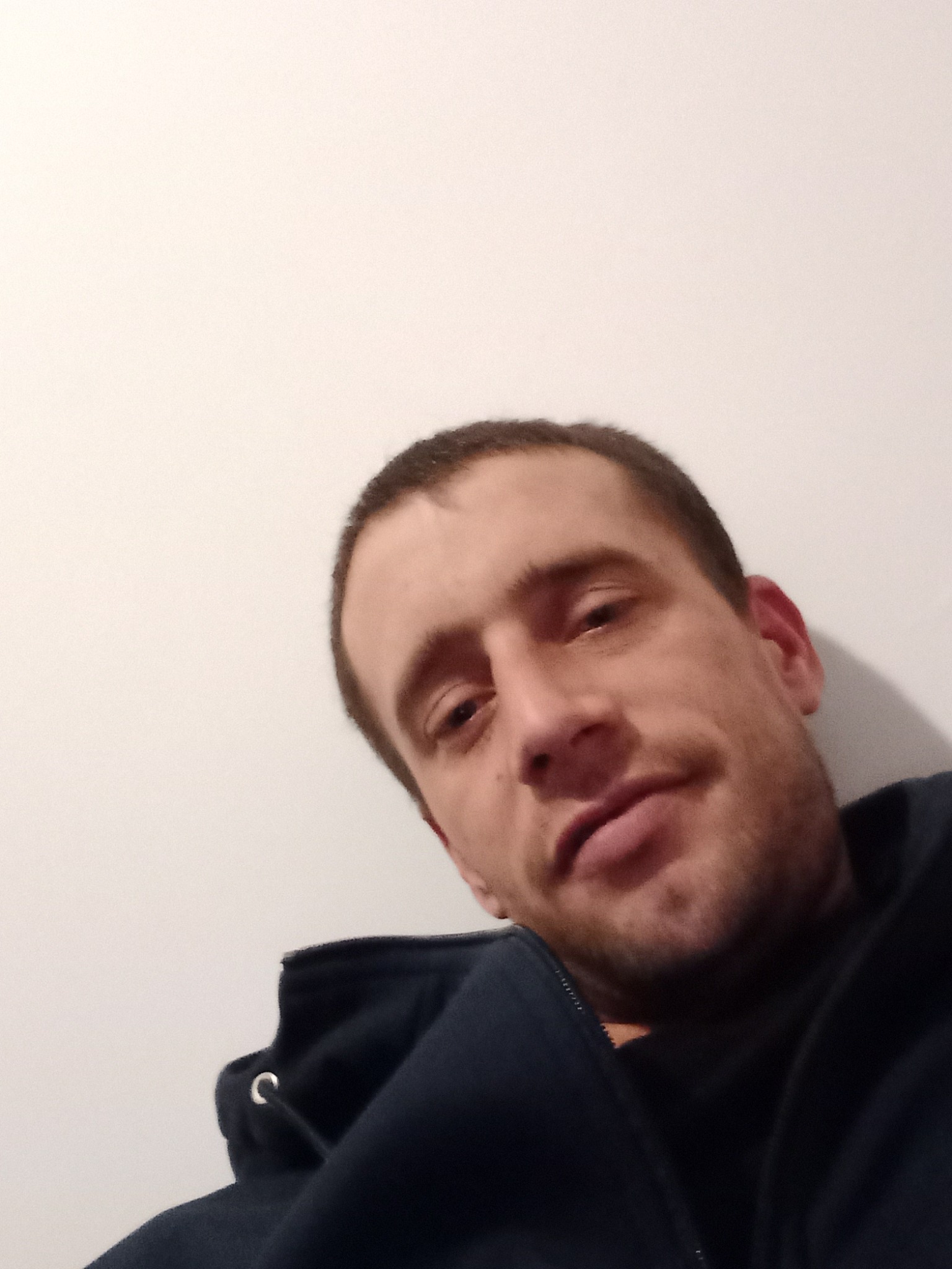 Portret mężczyzny z bliska, ubrany w ciemną bluzę z kapturem, na tle jasnej ściany, ujęcie z dołu.