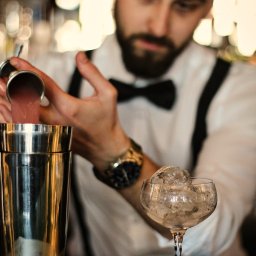 Barmates - Barman w białej koszuli z muchą i szelkami przelewa różowy płyn z miarki do shakera, obok stoi kieliszek wypełniony lodem, widoczny złoty zegarek na nadgarstku.