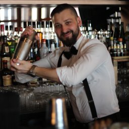 Barmates - Uśmiechnięty barman w białej koszuli z muchą i szelkami potrząsa shakerem za barem pełnym butelek alkoholu, widoczne szkło barowe.