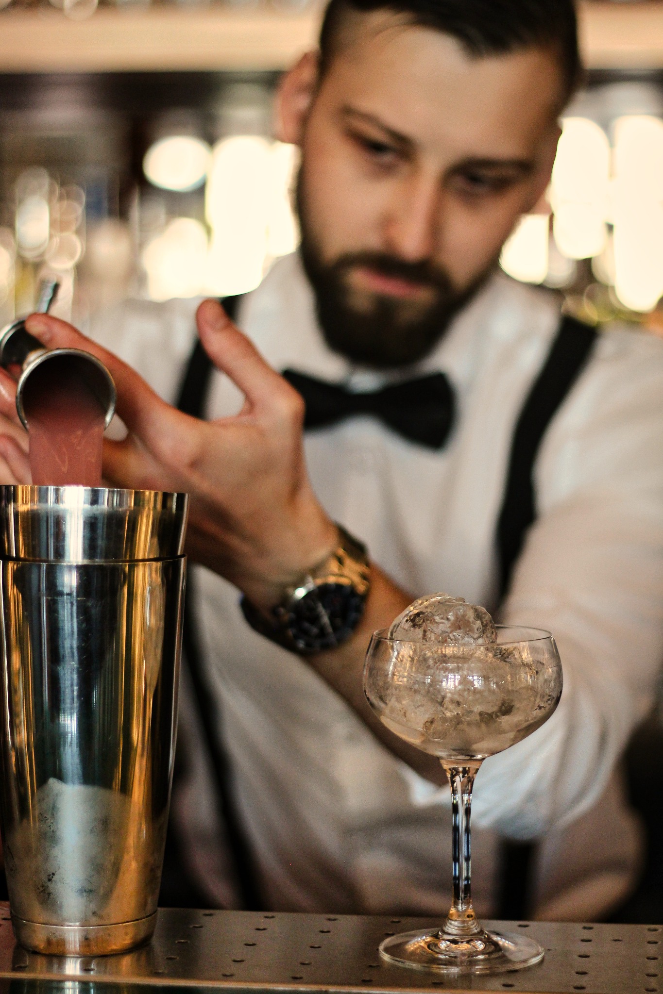 Barman w białej koszuli z muchą i szelkami przelewa różowy płyn z miarki do shakera, obok stoi kieliszek wypełniony lodem, widoczny złoty zegarek na nadgarstku.