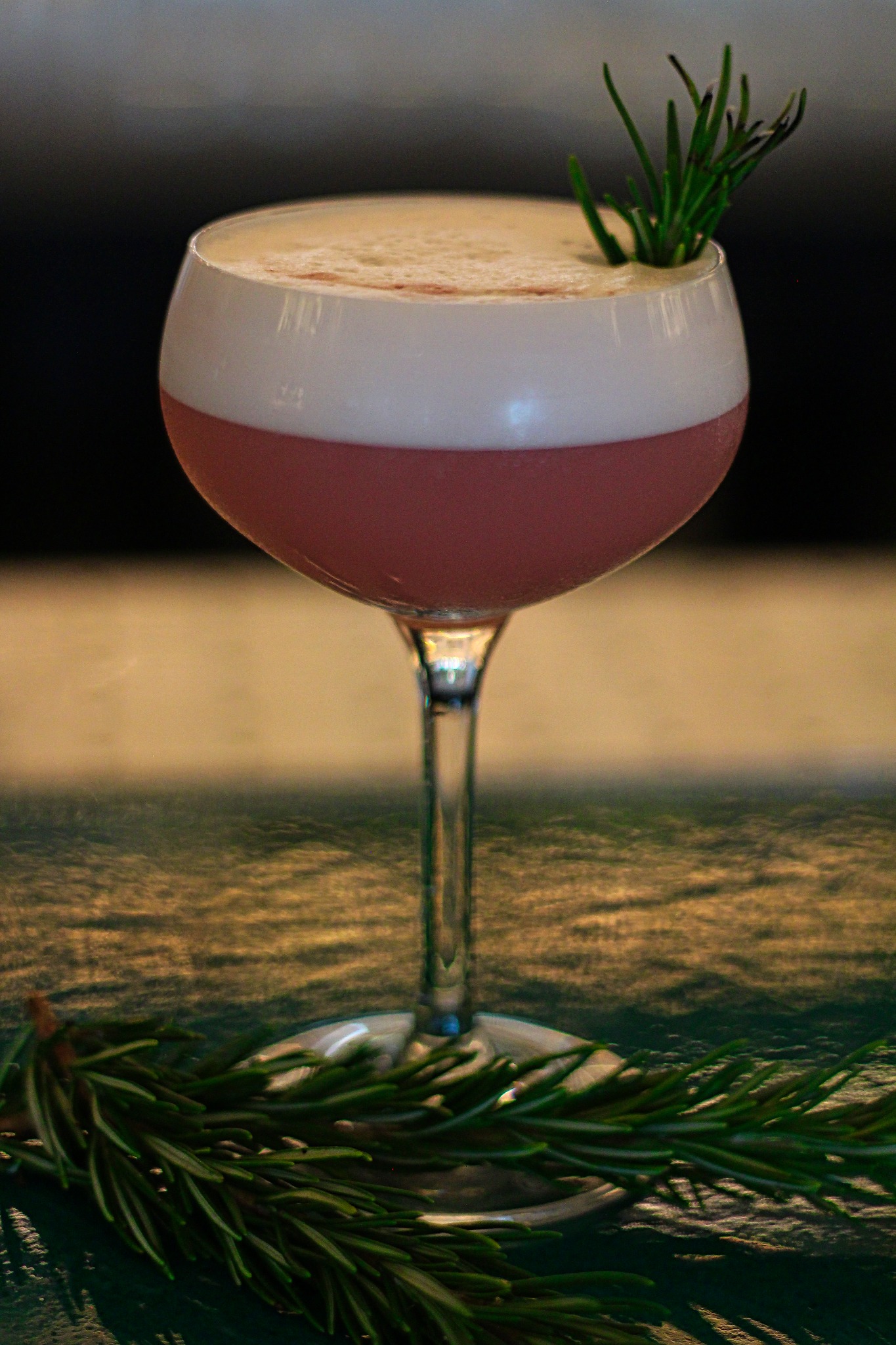Elegancki kieliszek z różowym koktajlem zwieńczonym pianką, udekorowany gałązką rozmarynu, leżącą również u podstawy kieliszka na teksturowanym blacie.