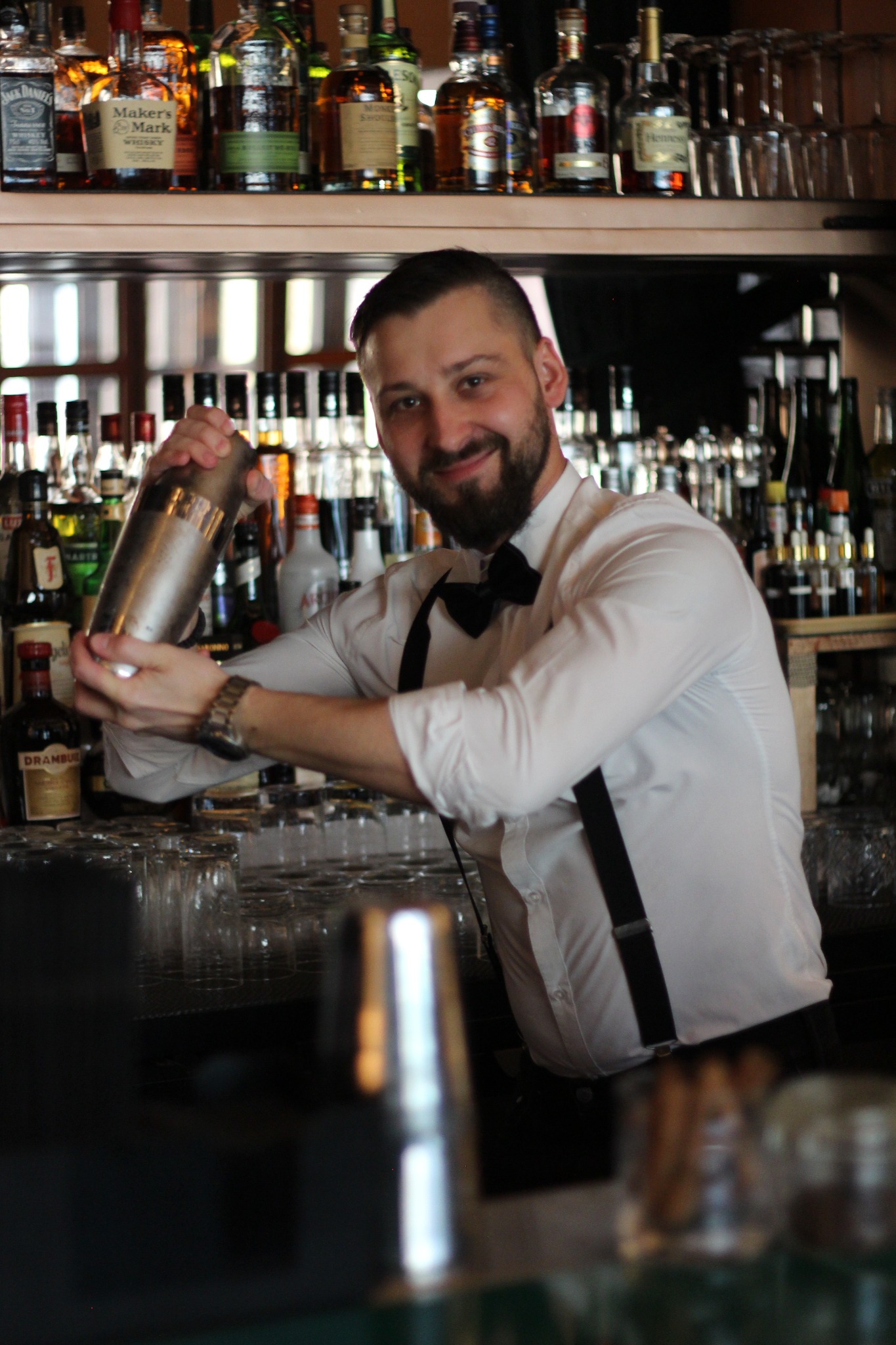 Uśmiechnięty barman w białej koszuli z muchą i szelkami potrząsa shakerem za barem pełnym butelek alkoholu, widoczne szkło barowe.