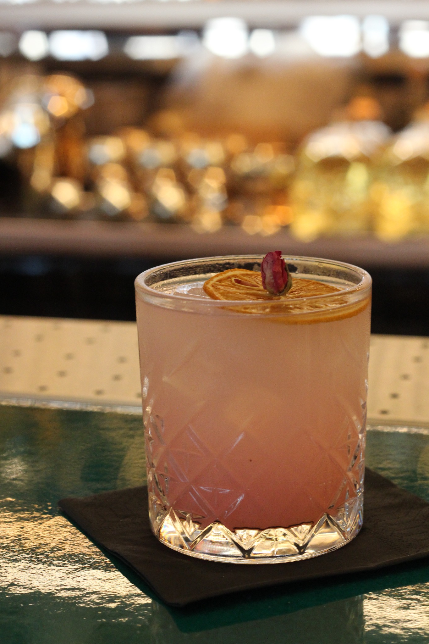 Różowy koktajl w szklance z dekoracją z suszonej pomarańczy i pączka róży, serwowany na czarnej serwetce, z rozmytym tłem baru.