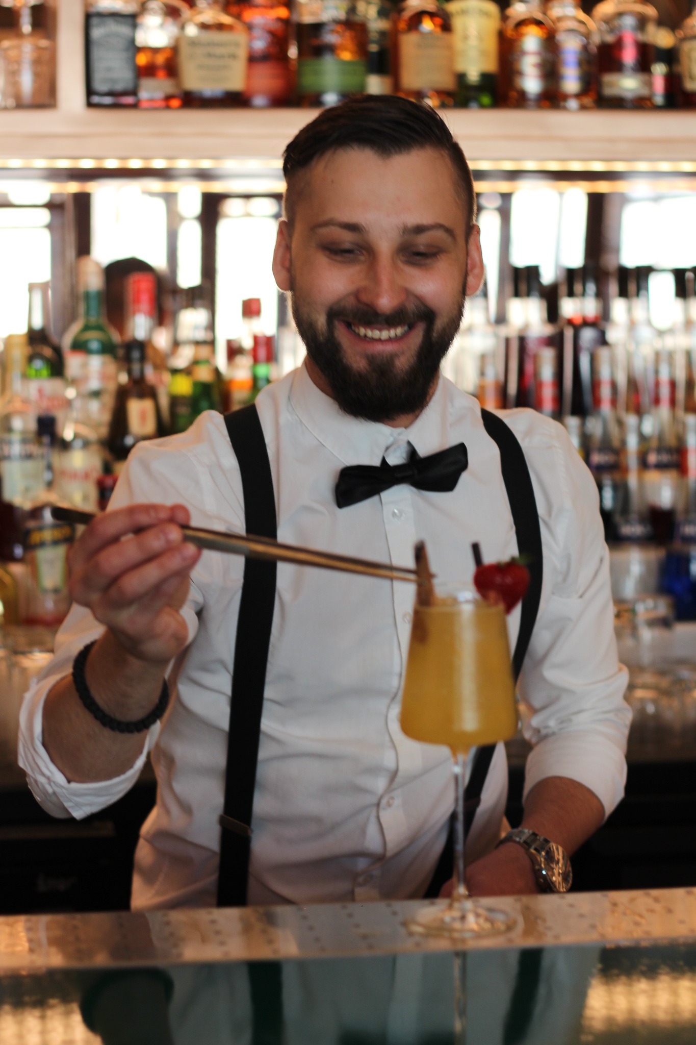 Uśmiechnięty barman w białej koszuli z muchą i szelkami przygotowuje koktajl z truskawką i laską cynamonu, używając długiej, metalowej łyżki barmańskiej, na tle półek z butelkami alkoholi.