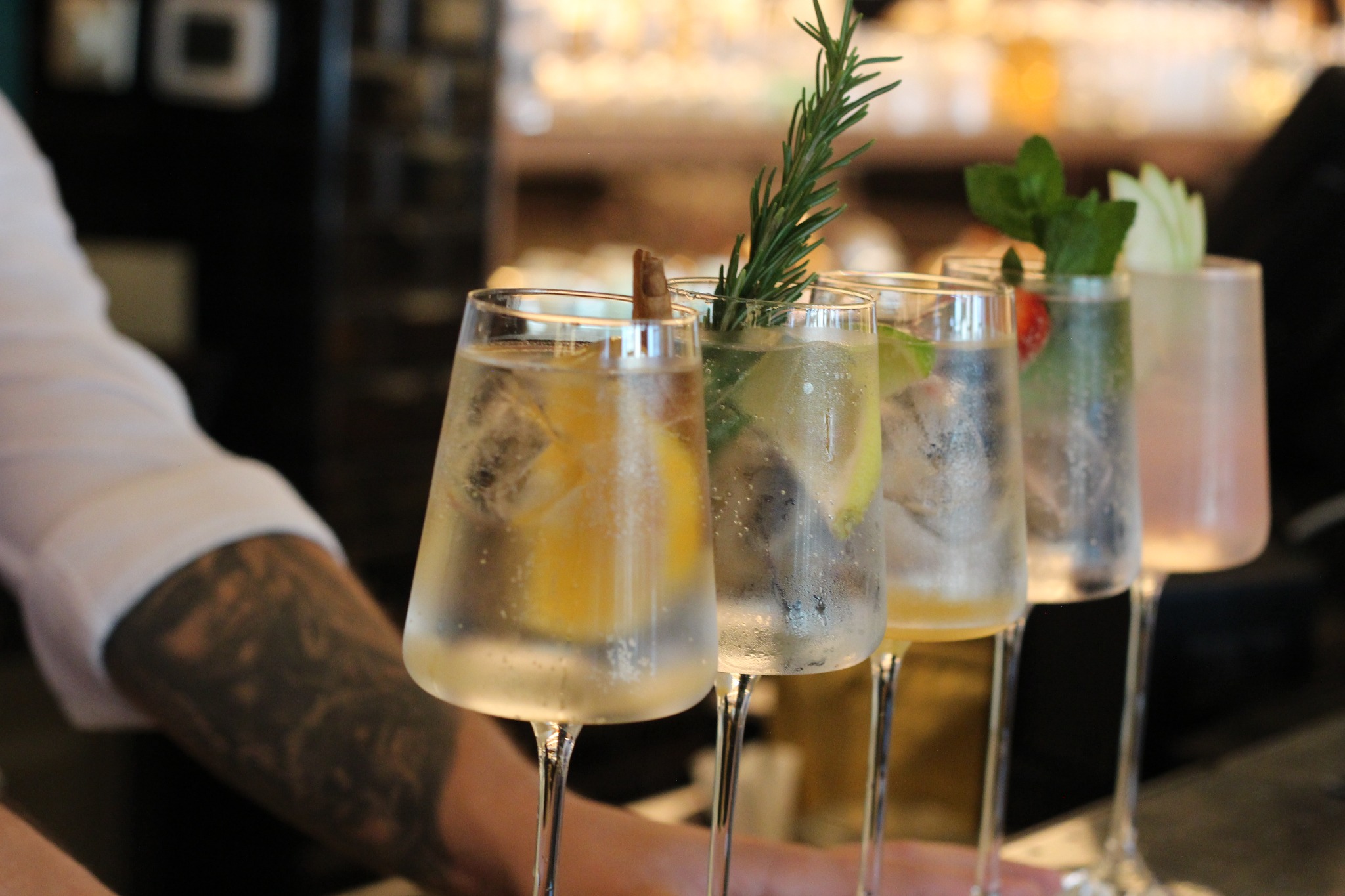 Cztery wysokie szklanki z orzeźwiającymi napojami na barze, każda udekorowana inaczej: plaster cytryny i laska cynamonu, gałązka rozmarynu, truskawka i mięta, oraz plaster ogórka. Widoczna...