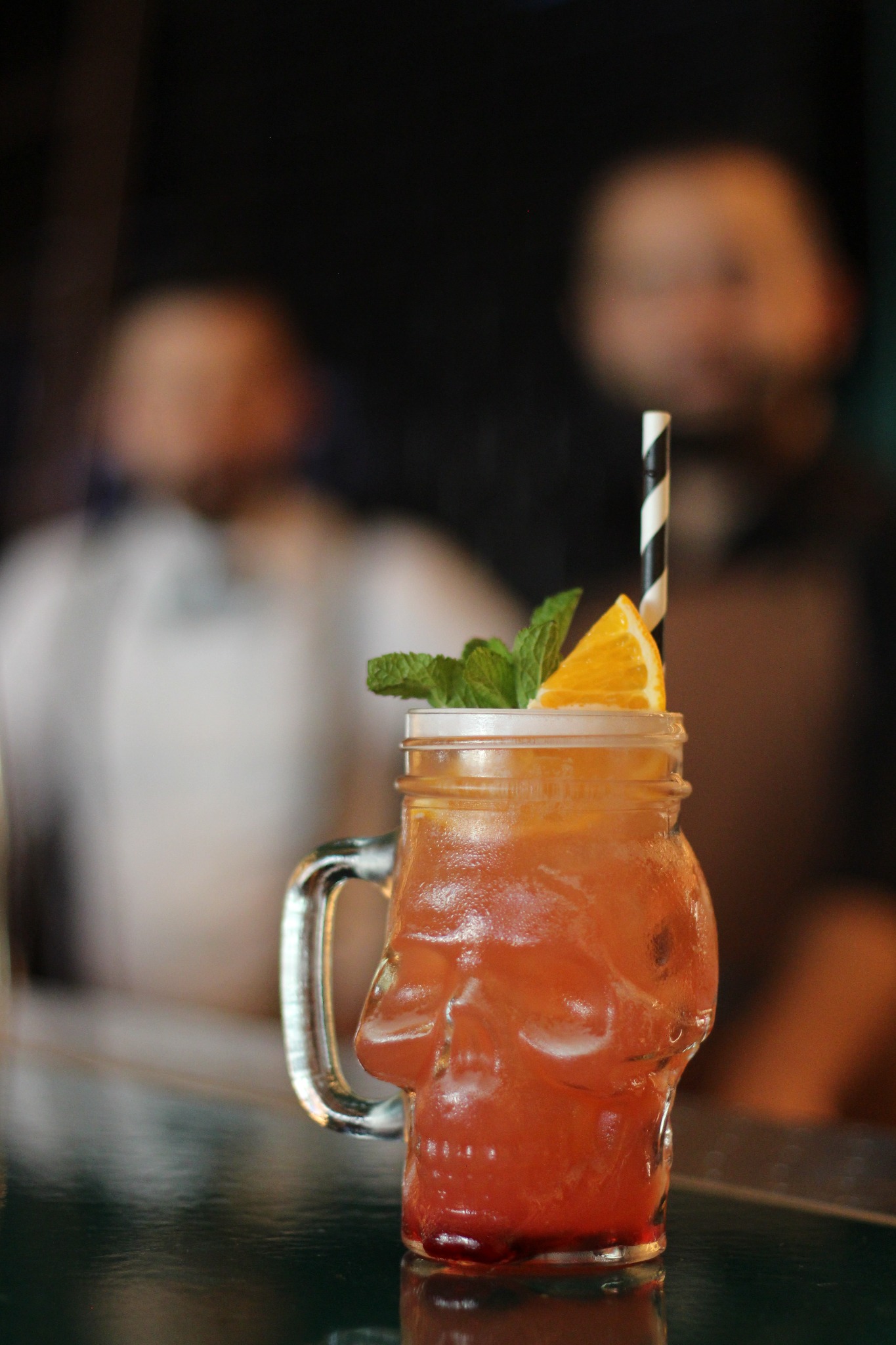 Pomarańczowy koktajl w szklance w kształcie czaszki, ozdobiony miętą i plasterkiem pomarańczy, z czarno-białą słomką, na błyszczącej powierzchni, z rozmytymi postaciami w tle.