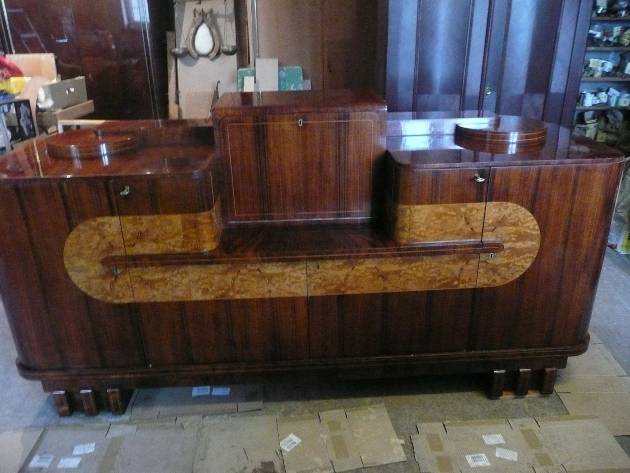 Odrestaurowany, lakierowany kredens art deco z fornirem orzechowym i intarsjami z jasnego drewna, widoczne ślady renowacji na podłodze.
