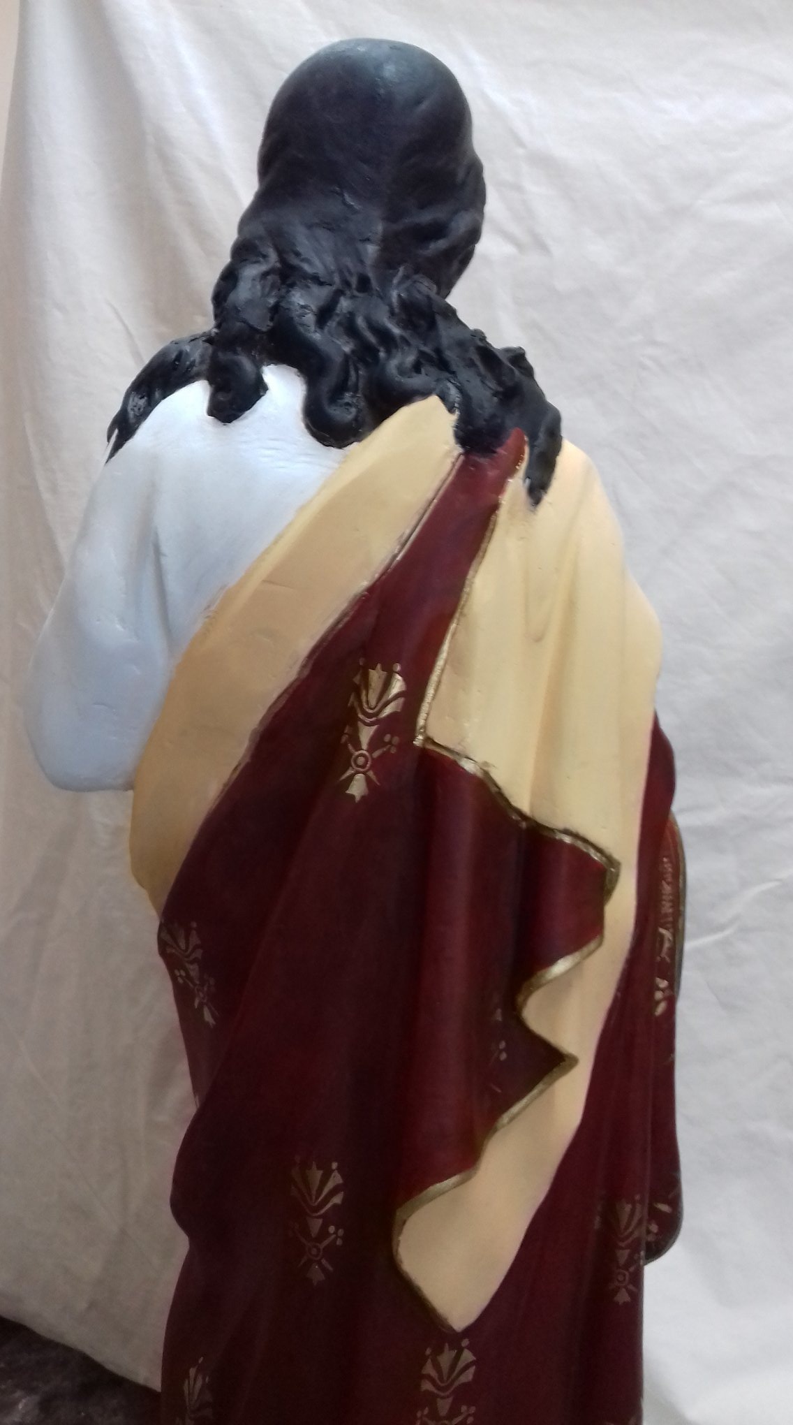 Tył figury religijnej z czarnymi kręconymi włosami i szatą w odcieniach czerwieni i beżu, zdobioną złotymi ornamentami.