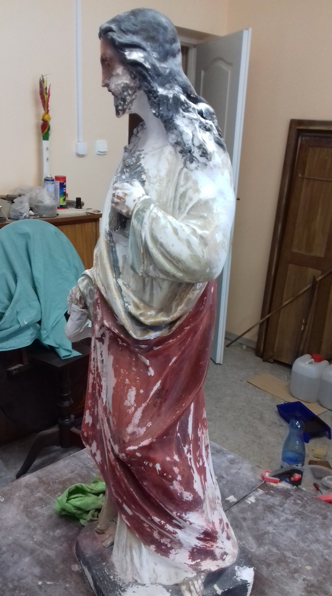 Figura sakralna w trakcie renowacji w Warszawie, widoczne ślady farby i materiały konserwatorskie na stole roboczym.