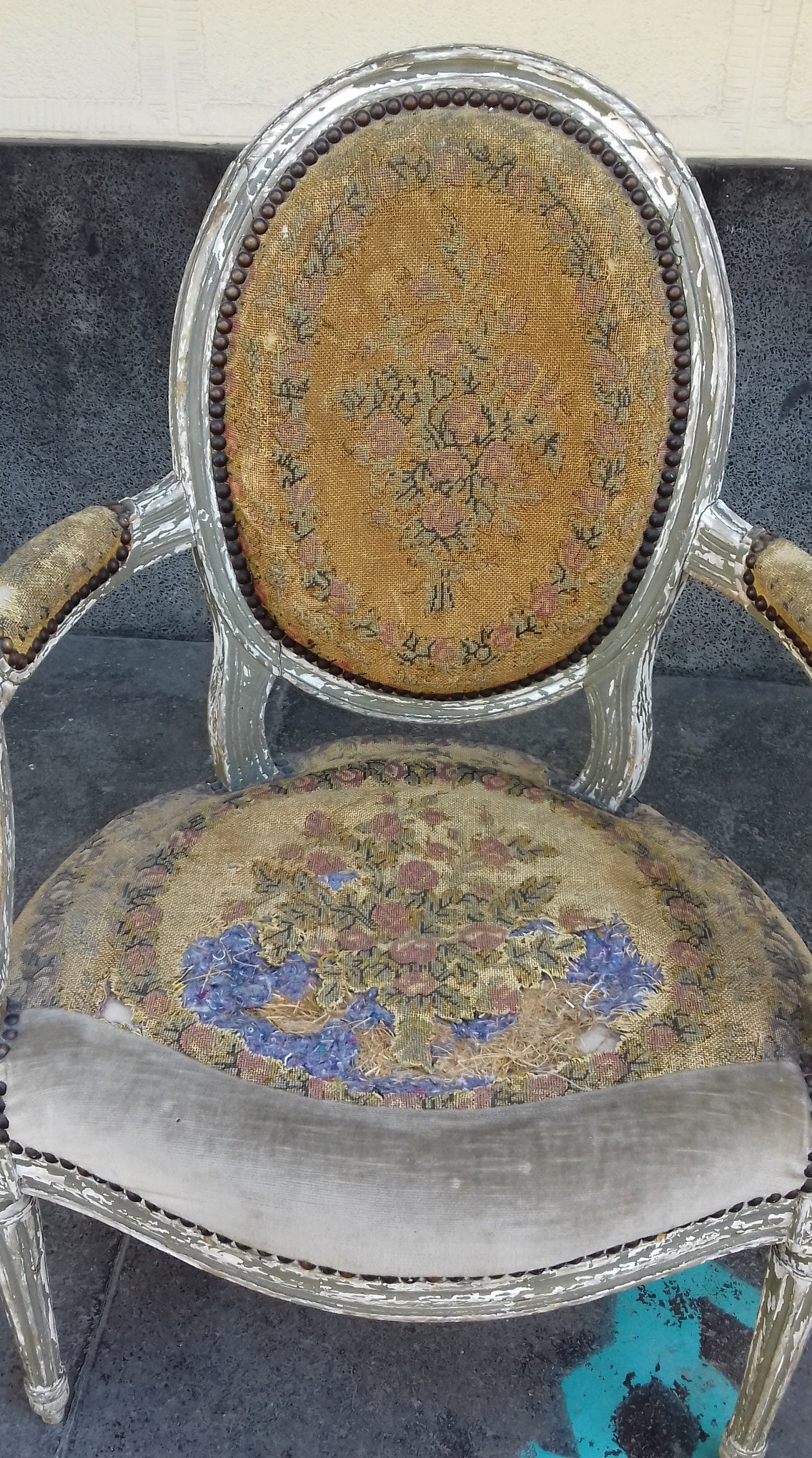 Antyczne krzesło z owalnym oparciem, zniszczona tapicerka z motywem kwiatowym, widoczne ślady użytkowania i odpryski farby na ramie.