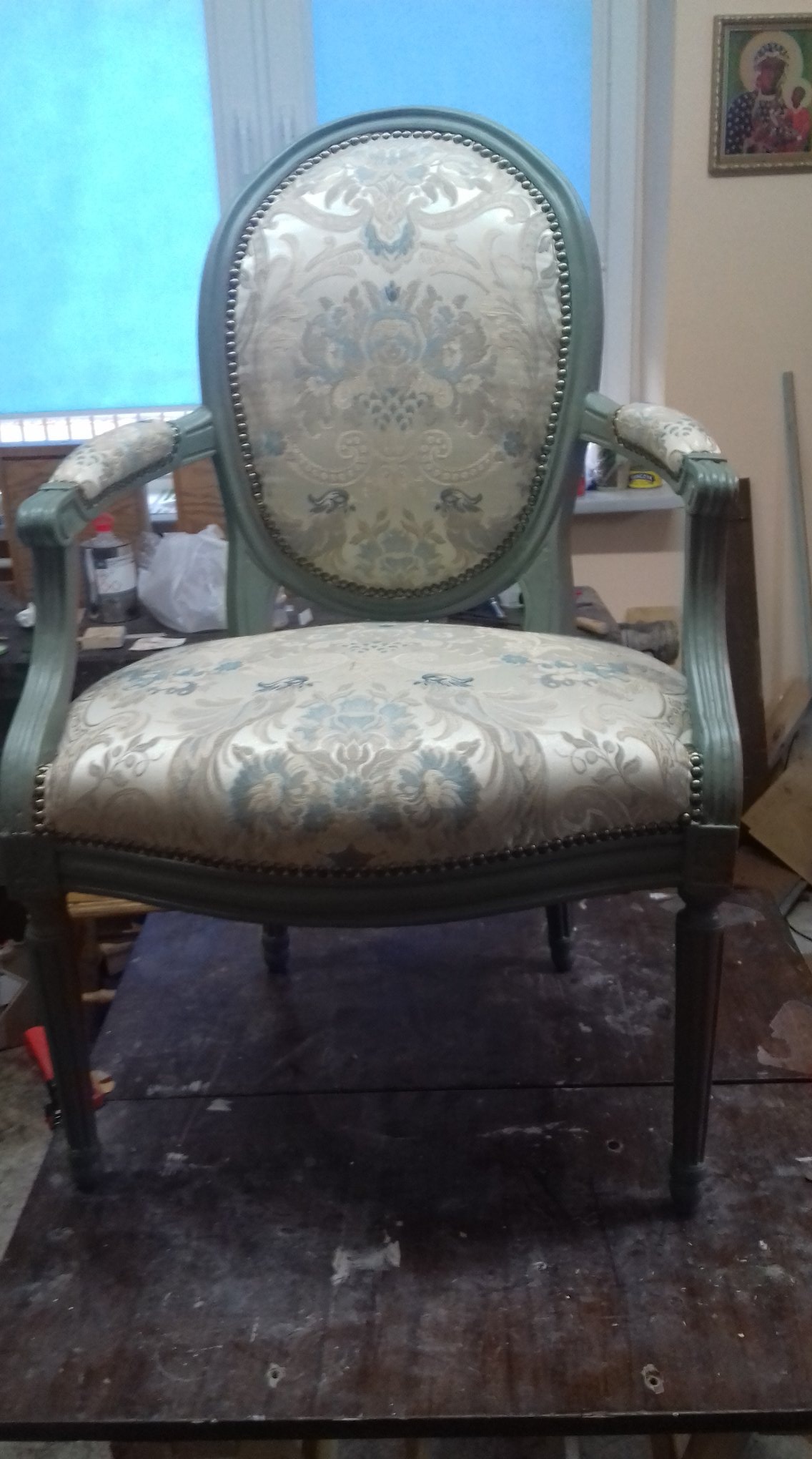 Odrestaurowane krzesło w stylu Ludwika XVI z owalnym oparciem, tapicerowane wzorzystą tkaniną w odcieniach beżu i błękitu, z ramą w kolorze szarozielonym, stojące na drewnianym stole warsztatowym.
